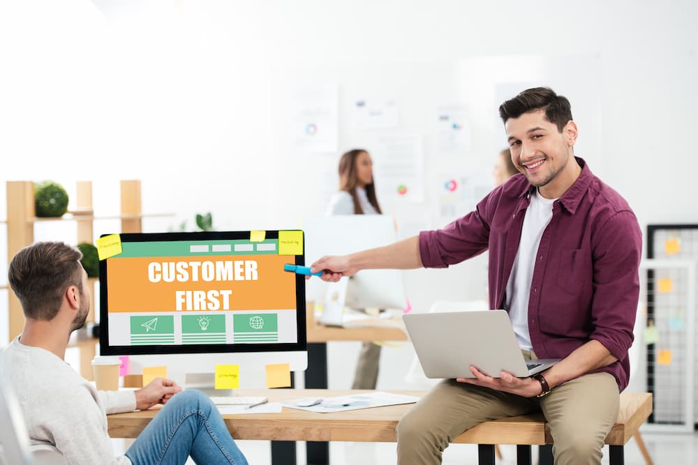 Two young adults are collaborating on sales and marketing approach, one pointing a pen at a monitor screen with the words "customer first".